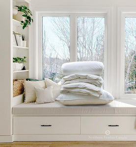 Folded Baltic Goose down Duvet (twin fill level 2) ontop of a Baltic Goose Down Pillow (medium fill) resting on a window bench beside white bookshelves filled with plants