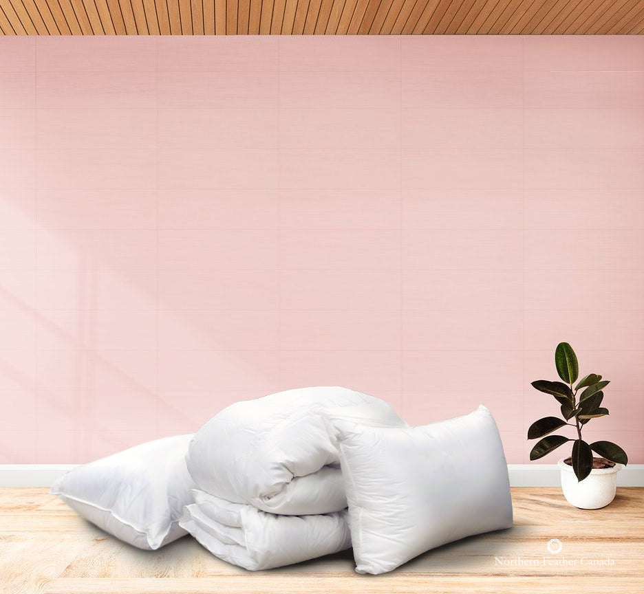 Folded Siberian Snow Goose Down Duvet (Twin sized with level 3 fill) with 2 Siberian Snow Goose Down Pillows (medium fill) on a hardwood floor with soft pink walls and a rubber plant in a white pot