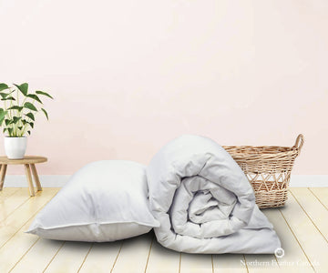 Rolled White Goose Down Duvet (fill level 2) and White Goose Down Pillow (Medium fill) on maple hardwood flooring with wicker basket and plant in background